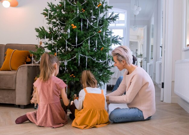 Mamá e hijas decoran un árbol de Navidad sentados en el suelo de la sala de estar