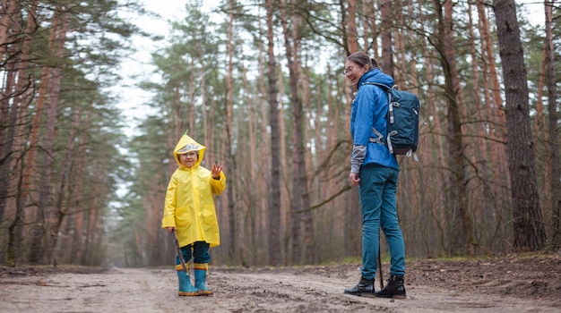 Mamá e hijo caminan por el camino forestal después de la lluvia en impermeables juntos