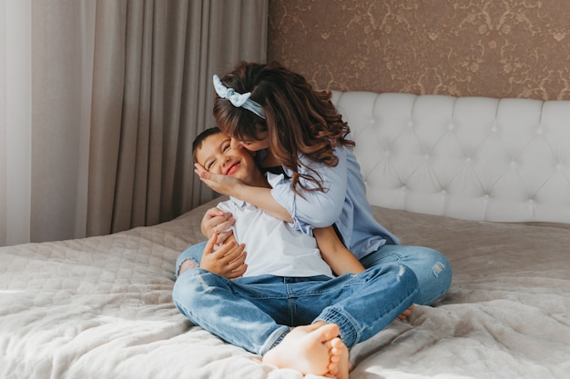 Mamá e hijo están sentados en la cama, abrazándose y divirtiéndose. Amar a mamá e hijo juntos. ¡Feliz día de la madre!