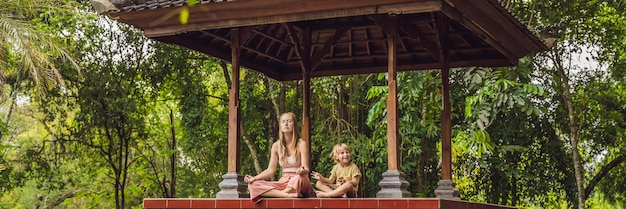 Mamá e hijo meditan practicando yoga en el tradicional cenador balinés BANNER, formato largo