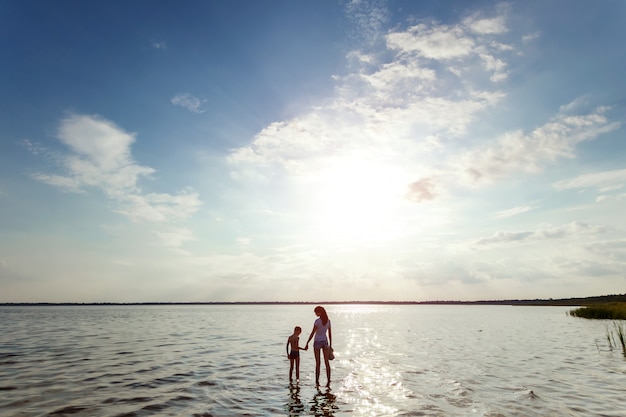 Mamá e hijo se paran en el agua contra la hermosa puesta de sol en el lago.