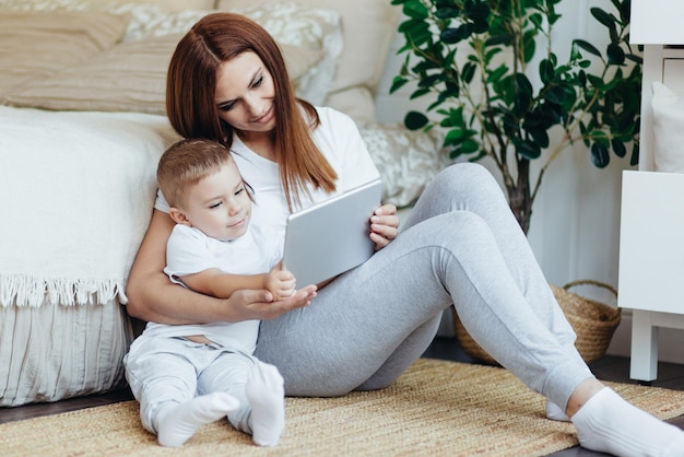 Mamá e hijo se sientan en el suelo de la habitación y miran juntos la tableta digital