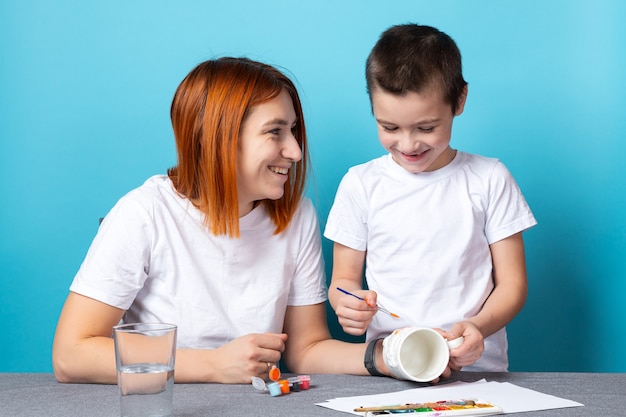 Mamá e hijo sonríen alegremente y pintan la tapa en naranja brillante sobre fondo azul. Concepto de aprendizaje de dibujo para niños