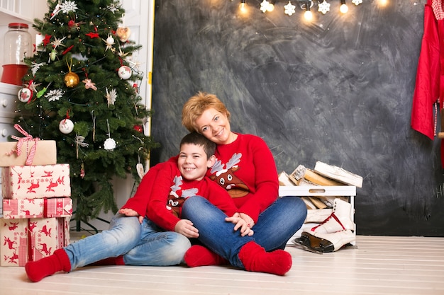 Mamá e hijo en suéteres rojos de Navidad, divirtiéndose en el árbol de Navidad, abrazándose y riendo