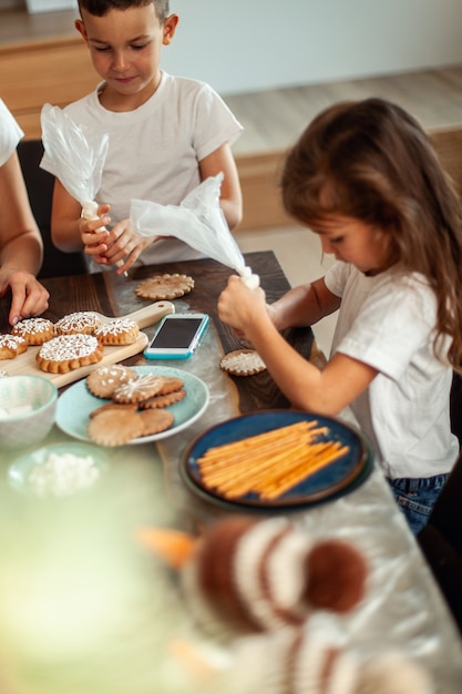 Mamá e hijos decoran pan de jengibre navideño en casa. Un niño y una niña pintan con cornetas con