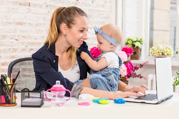 Mamá y empresaria trabajando con computadora portátil en casa y jugando con su bebé.