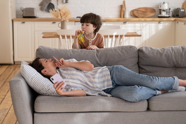 Mamá estresada le pide a su hijo activo que calme a la madre cansada, intente descansar después del trabajo en el sofá, sostenga el teléfono inteligente