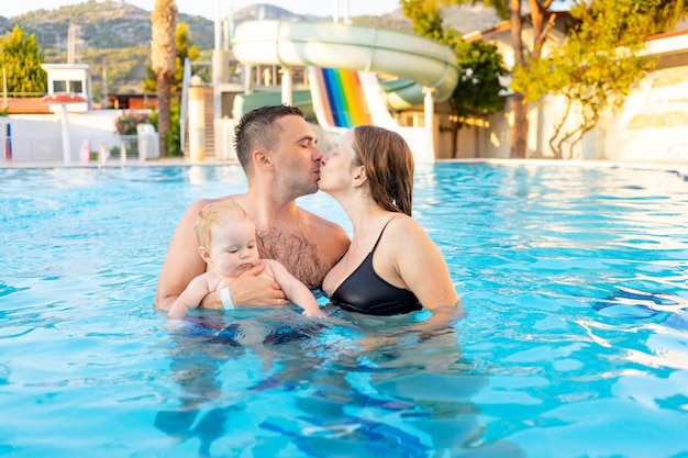 Mamá de familia feliz, papá e hija están nadando en la piscina con toboganes de agua y divirtiéndose en las vacaciones, besándose y abrazándose