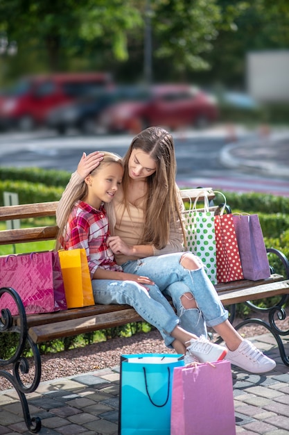 Mamá feliz abrazando a su hija, sentada en un banco en el parque después de ir de compras