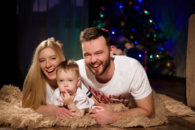 Mamá feliz alegre, papá e hijo pequeño de 5 años en el fondo del árbol decorado antes de Navidad se abrazan. Ambiente acogedor navideño.