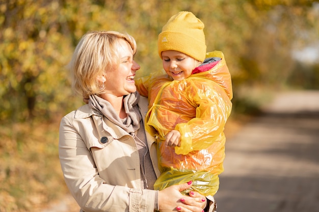 Mamá feliz joven sostiene a la niña con sombrero amarillo e impermeable en sus brazos, sonriendo felizmente en el parque de otoño entre árboles amarillos