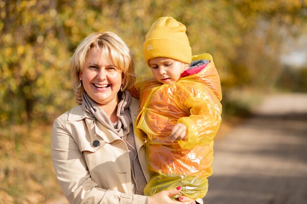 Mamá feliz joven sostiene a la niña con sombrero amarillo e impermeable en sus brazos, sonriendo felizmente en el parque de otoño entre árboles amarillos