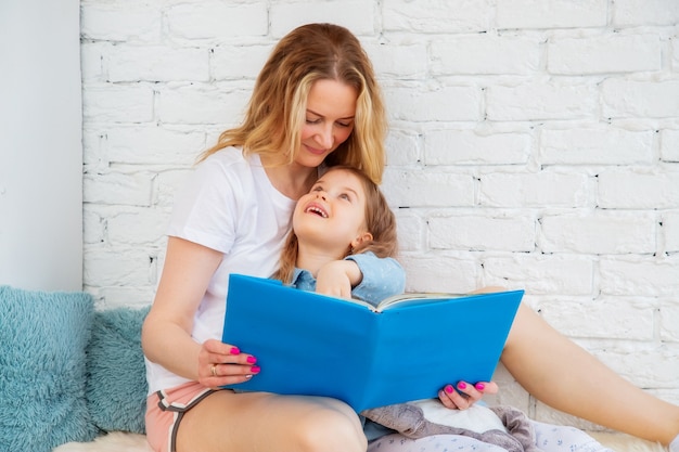 Mamá feliz lee un cuento de hadas alegre en un libro a su hija y sonríe mientras está sentada en la sala de estar