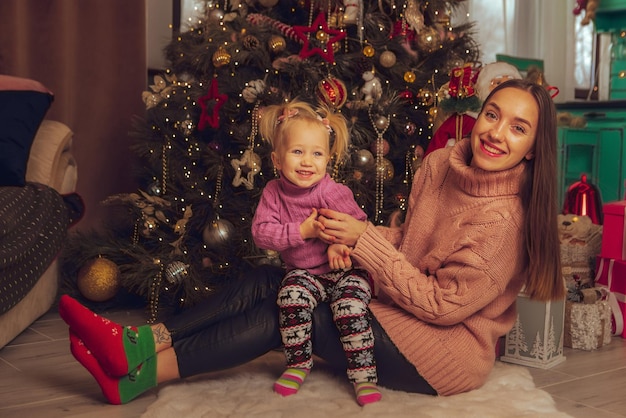 Mamá feliz y su pequeña niña sonríe en el árbol de navidad