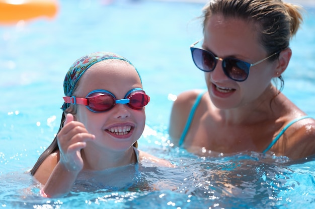 Mamá en gafas de sol enseñando a su hija a nadar en la piscina