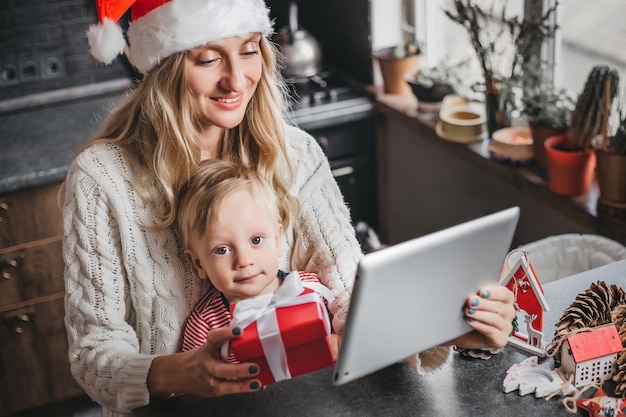 Mamá con un gorro de Papá Noel se sienta con un niño en casa, sostiene una tableta y hace una videoconferencia.