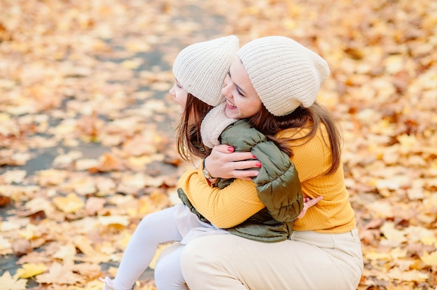 Mamá con un gorro de punto mira suavemente a su hija de cinco años sosteniéndola en sus brazos en el otoño contra el fondo de hojas amarillas caídas