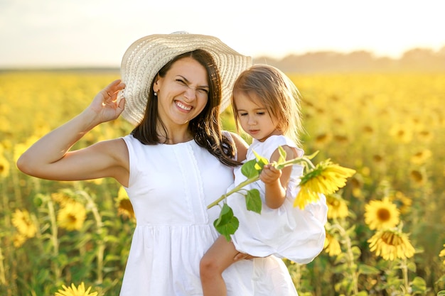 Mamá hace una mueca graciosa riéndose alegremente y sostiene a la niña en brazos en el campo con girasoles en crecimiento