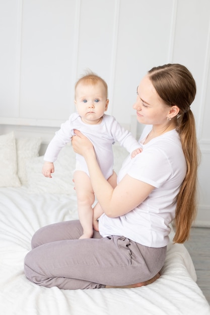 Mama hält Baby in ihren Armen und umarmt ihn sanft auf dem weißen Bett zu Hause, Mutterliebe