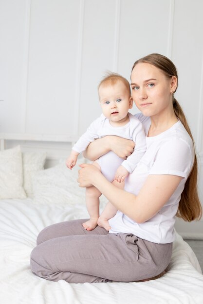 Mama hält Baby in ihren Armen und umarmt ihn sanft auf dem weißen Bett zu Hause, Mutterliebe.