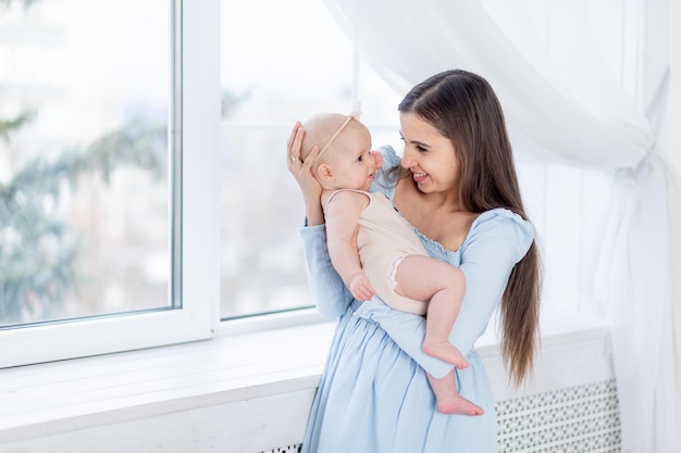 Mama hält ein neugeborenes Mädchen in einem Baumwollanzug am Fenster des Hauses, umarmt und küsst ihn glückliche Mutterschaft oder Familie