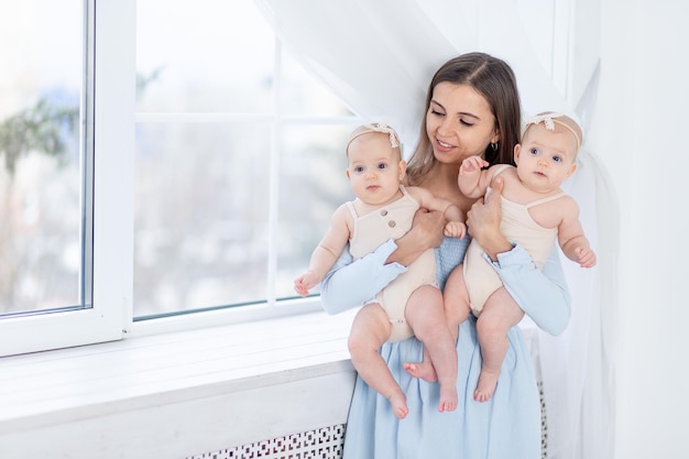 Mama hält zwei neugeborene Zwillingsmädchen in einem Baumwollanzug am Fenster des Hauses, umarmt und küsst sie, Mutterschaft mit zwei Kindern