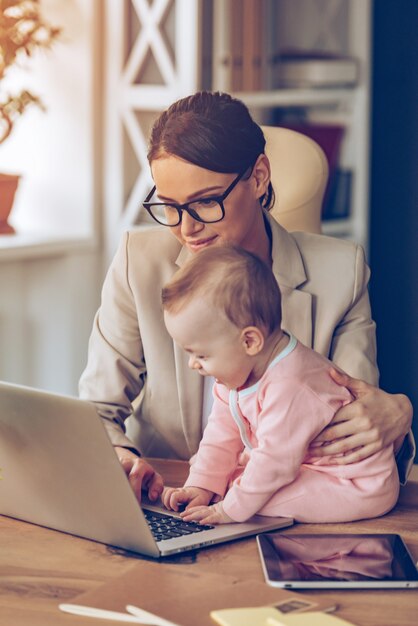 Mama helfen. Kleines Mädchen mit Laptop beim Sitzen auf dem Schreibtisch mit ihrer Mutter im Büro