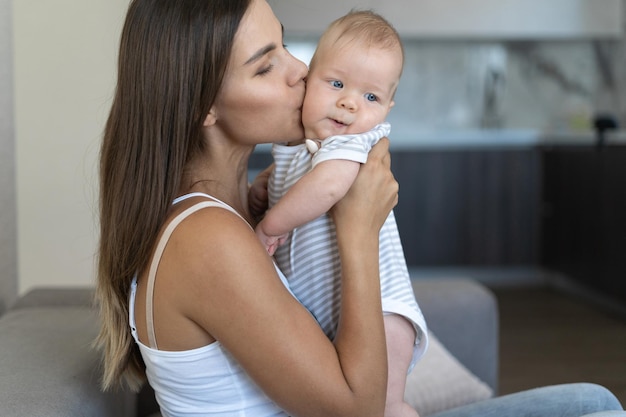 Mamá hermosa joven está sentada en el sofá. Sostiene a un bebé recién nacido en sus brazos. besar a un niño en la mejilla