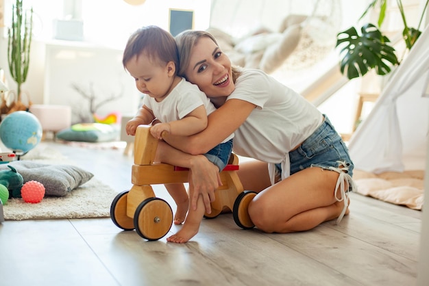 Mamá y hija pequeña pasan tiempo en la sala de juegos Niña pequeña en un scooter de madera Concepto de amor de cuidado materno