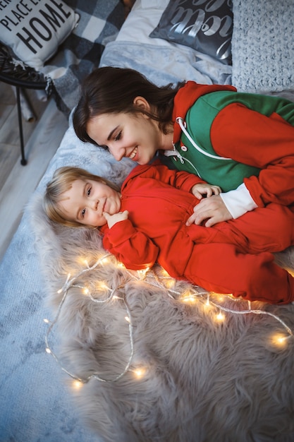 Mamá con hija en suéteres rojos calientes están saltando sobre la cama. Feliz maternidad. Relaciones familiares cálidas. Interior de Navidad y año nuevo. Amor. Concepto de familia.