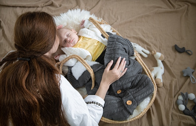 Mamá se inclinó sobre un bebé dormido en una cuna de mimbre con una vista superior de una manta de punto cálido.