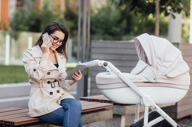 Mamá independiente exitosa que trabaja con el teléfono, sentada en un banco cerca del cochecito de bebé en el parque.