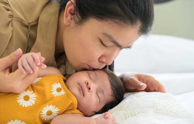 Mamá joven asiática besando a su linda niña recién nacida durmiendo en una cama blanca en casa Protección del amor de la atención médica y concepto del día de la madre