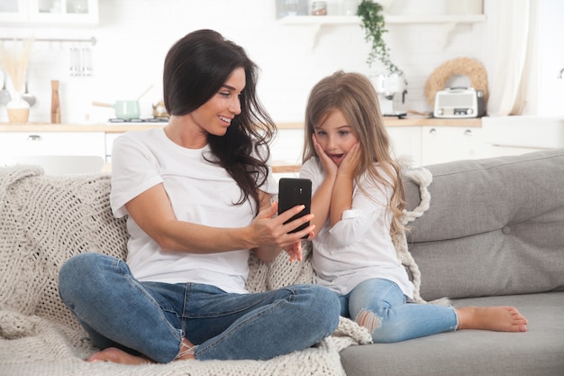 Mamá joven e hija pequeña jugando juegos en línea en casa usando un teléfono inteligente