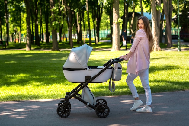 Mamá joven elegante camina con el cochecito en el parque