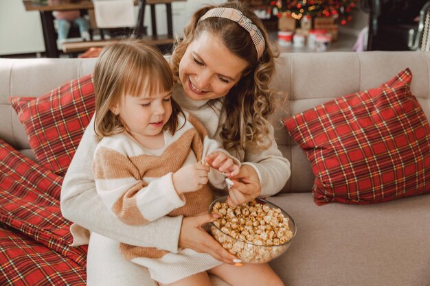 Mamá joven feliz y niña ríen mientras están sentados en el sofá y comen palomitas de maíz. familia viendo televisión y comiendo palomitas de maíz.