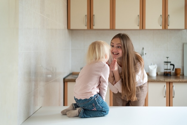 Mamá joven feliz y pequeña hija riendo en la cocina.