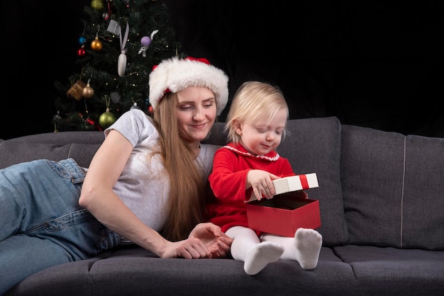 Mamá joven con gorro de Papá Noel y bebé desempaqueta el regalo de Navidad sobre fondo negro. Año nuevo.