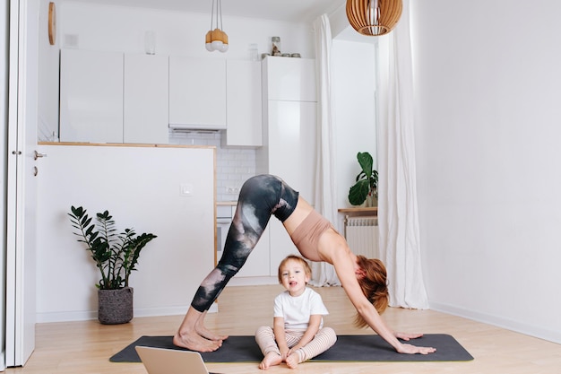 Mamá joven haciendo yoga junto a su hijo pequeño en la cocina de casa