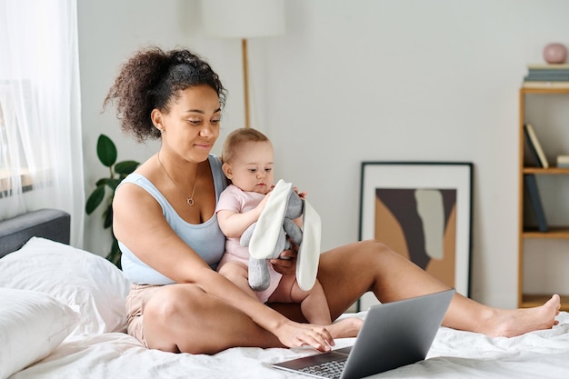 Mamá joven sentada en la cama con su bebé y usando una computadora portátil para su trabajo en línea