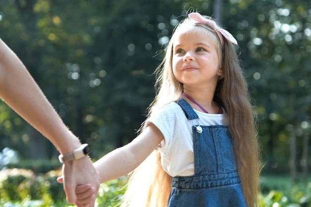 Mamá joven y su pequeña hija con cabello largo caminando juntos tomados de la mano en el parque de verano.