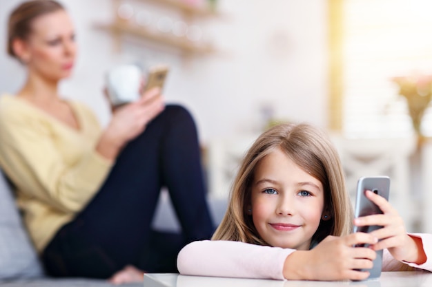 Mamá joven y su pequeña hija con smartphone y sentada en el sofá en casa