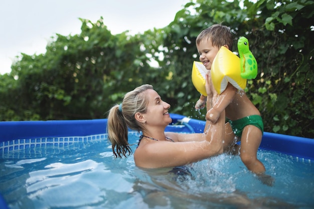 Mamá juega con un bebé desnudo en mangas largas en la piscina en el contexto de una puesta de sol de verano