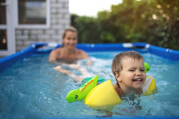 Mamá juega con un bebé desnudo en mangas en la piscina con el telón de fondo de una puesta de sol de verano