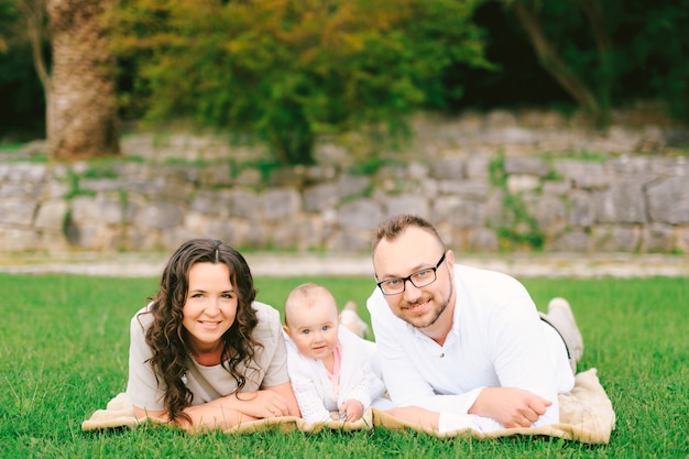 Mama, kleine Tochter und Papa liegen auf einer Decke auf einem grünen Rasen im Park vor dem Hintergrund von a