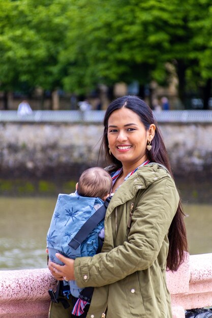 Mamá latina con su bebé en un portabebés azul feliz con los brazos abiertos en la playa y en primavera