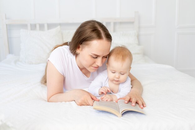 Mamá lee un libro al bebé antes de acostarse en una cama blanca con ropa de cama de algodón en casa, familia feliz