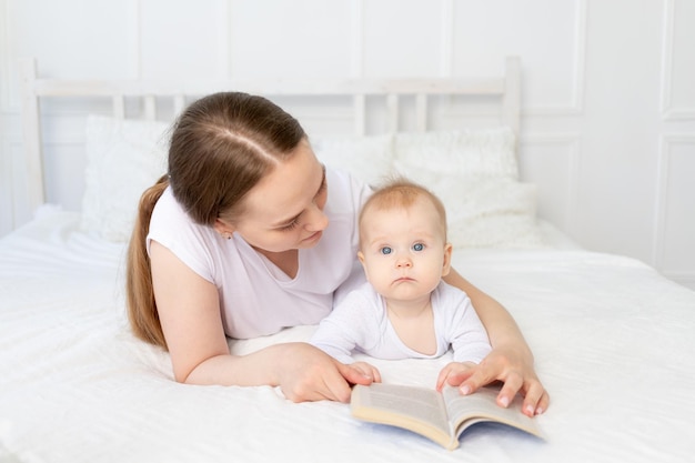 Mamá lee un libro al bebé antes de acostarse en una cama blanca con ropa de cama de algodón en casa familia feliz