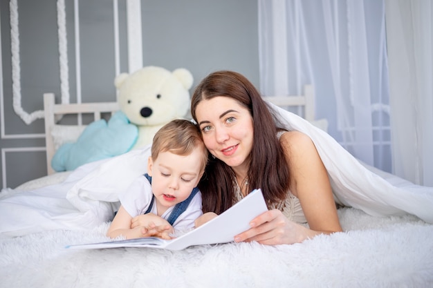 Mama liest dem Kind ein Buch vor oder bringt es zu Hause auf dem Bett bei. Glückliche, liebevolle Familie. Mutter und Sohn