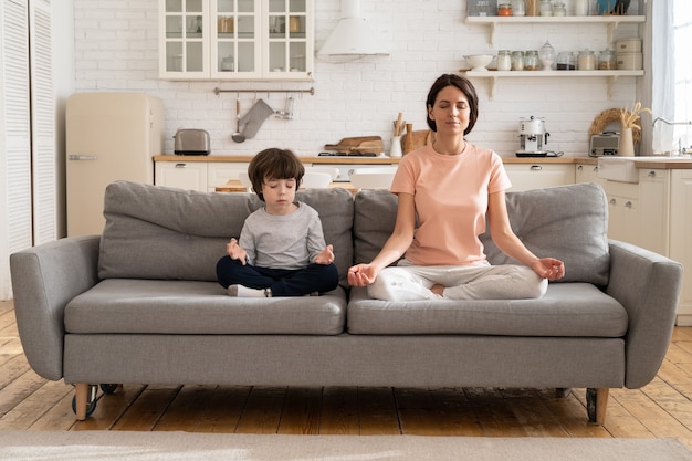 Mamá con lindo hijo pequeño haciendo ejercicio de yoga sentarse juntos en el sofá en casa enseñando al niño a meditar